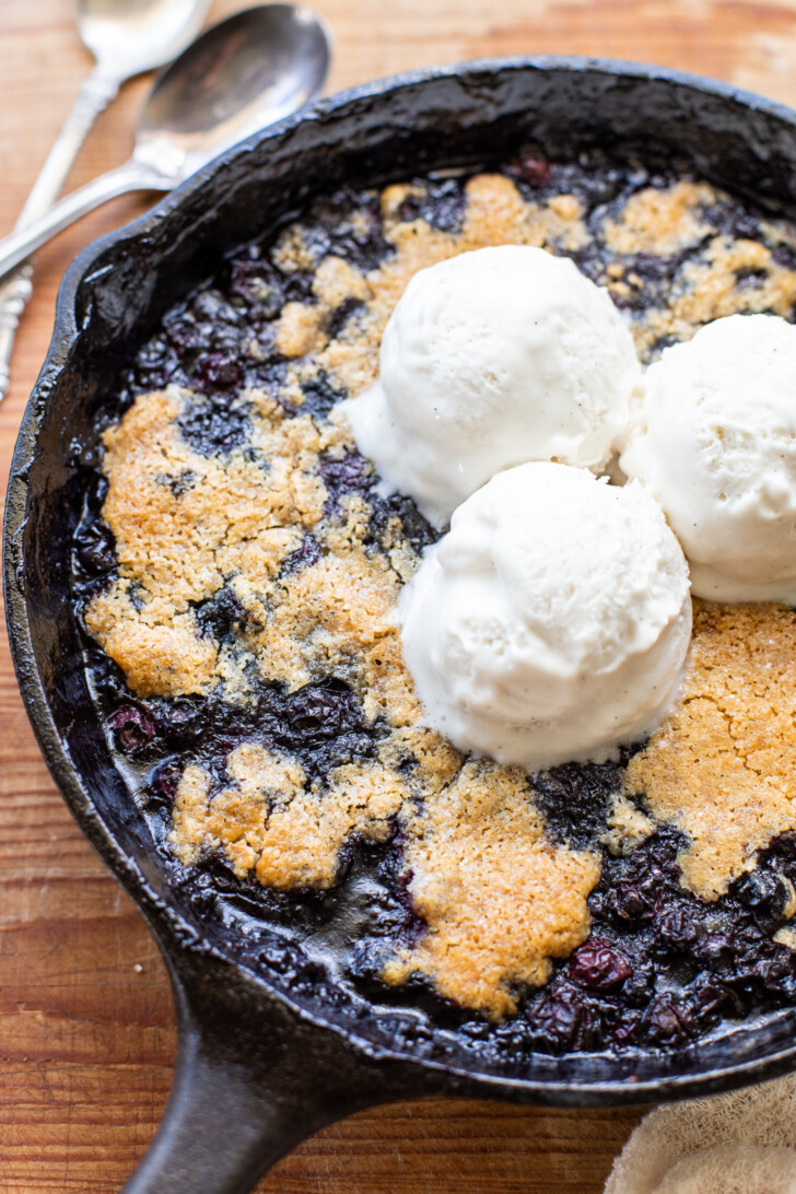 Gluten-free vegan blueberry skillet in a black cast iron skillet topped with three scoops of vegan vanilla ice cream, with a silver spoon on the top left.