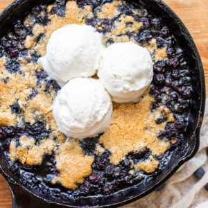 Gluten-free vegan blueberry skillet in a black cast iron skillet topped with three scoops of vegan vanilla ice cream, with a white napkin in the bottom right corner on a vintage wooden bread board.