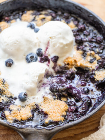 Gluten-free vegan blueberry skillet in a black cast iron skillet topped with three scoops of vegan vanilla ice cream. It has a spoonful taken out on the bottom right side with a white napkin in the bottom right corner on a vintage wooden bread board.