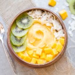 Kiwi slices, mango chunks, chia seeds, and coconut flakes topping a Mango Pineapple Smoothie Bowl in a wooden bowl with a silver spoon.