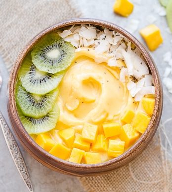 Kiwi slices, mango chunks, chia seeds, and coconut flakes topping a Mango Pineapple Smoothie Bowl in a wooden bowl with a silver spoon.