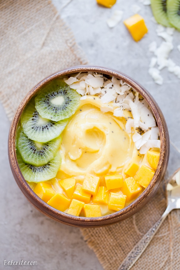 Kiwi, coconut, and mango chunks on top of a banana, mango, and pineapple smoothie bowl. On top of sheet pan with spoon, in a wooden bowl.