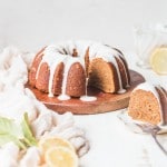 Gluten-Free Vegan Lemon Cake on wooden plate, with one slice taken out. Slice is in the front on a plate, with vegan lemon glaze and lemon garnish.