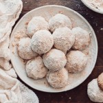A plate of round snowball cookies covered in powdered sugar sits on a wooden surface, surrounded by a white textured cloth. The cookies are arranged closely together, creating a cozy and inviting presentation.