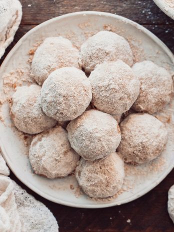 A plate of round snowball cookies covered in powdered sugar sits on a wooden surface, surrounded by a white textured cloth. The cookies are arranged closely together, creating a cozy and inviting presentation.