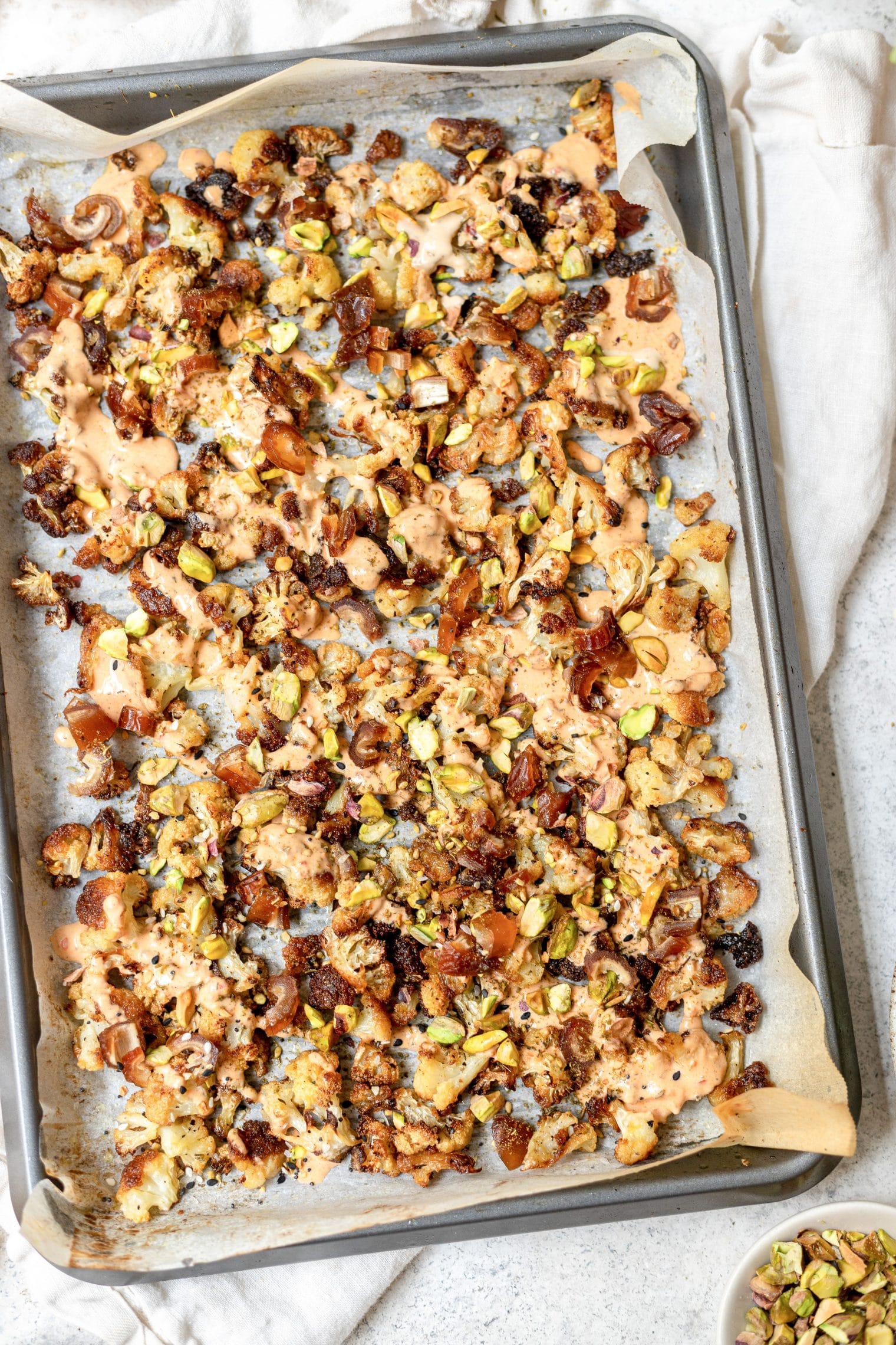 Cauliflower on a baking sheet on a white background. The cauliflower is drizzled with tahini harissa sauce, and topped with pistachios and Medjool dates.