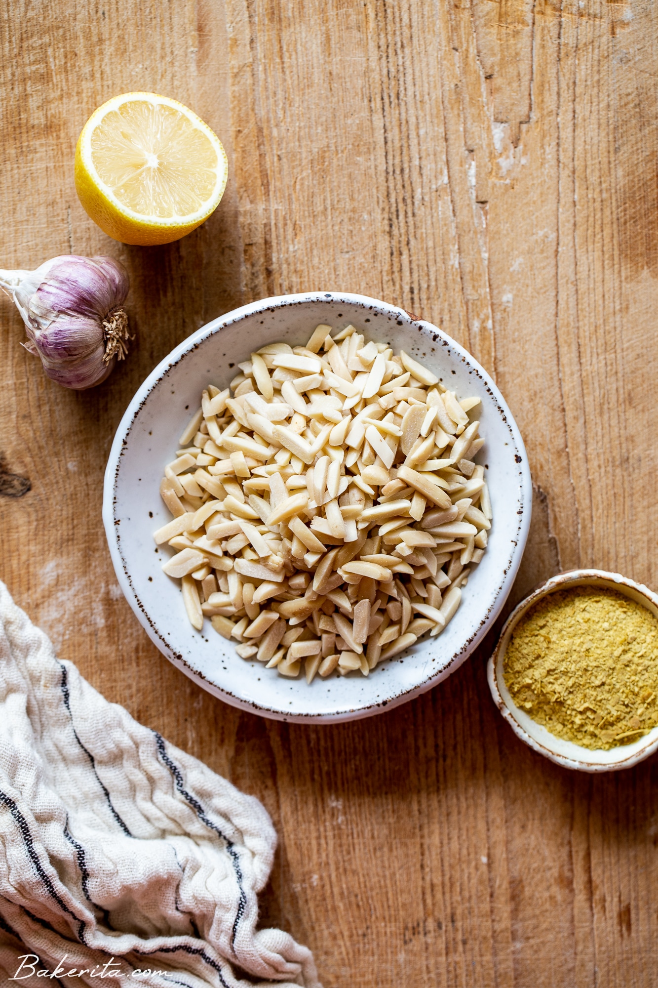 Wood background with slivered almonds in a white ceramic bowl, nutritional yeast, garlic, and lemon around the board.