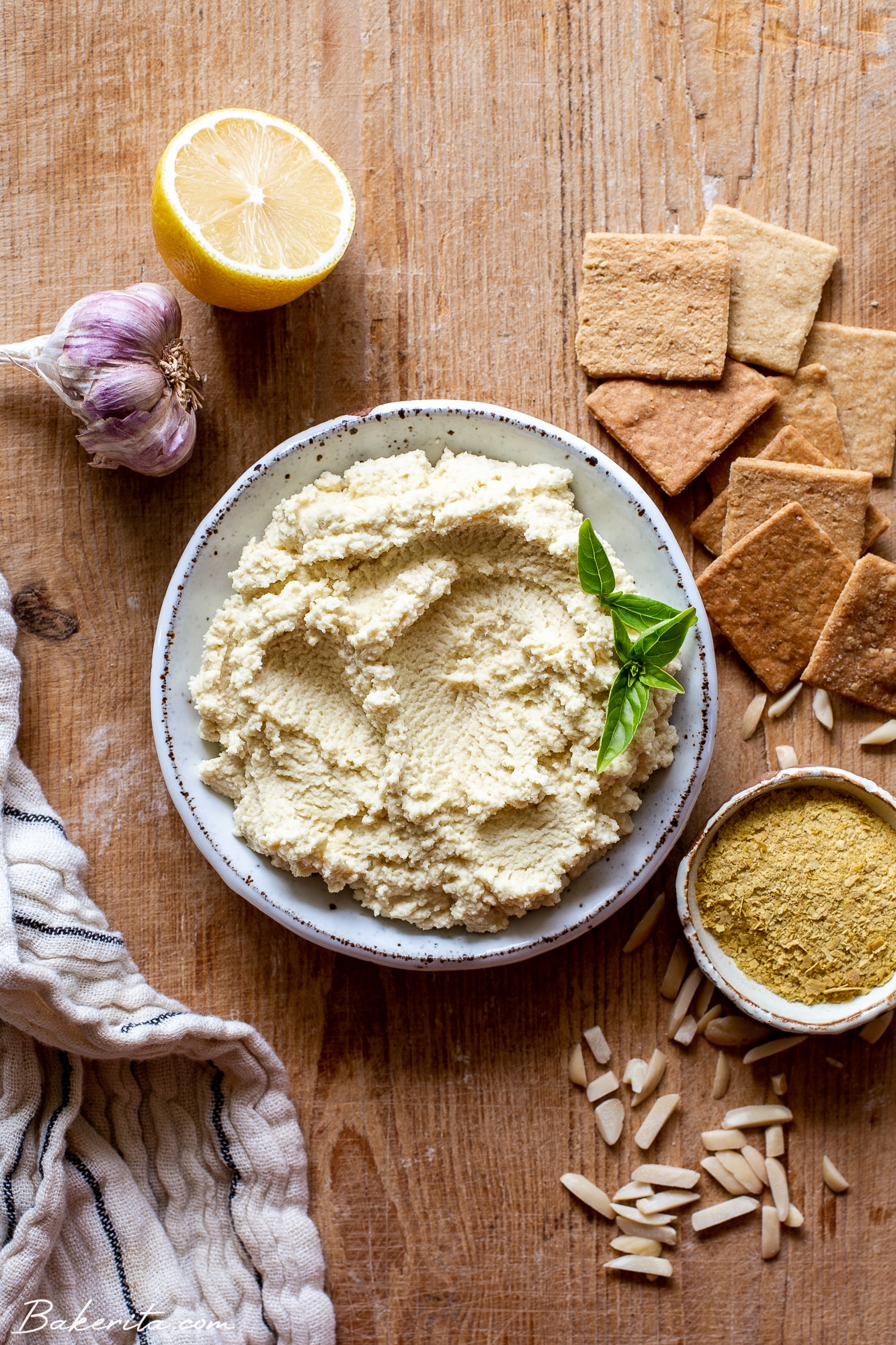 White ceramic bowl filled with vegan ricotta, garnished with basil leaves. On a wood background with garlic, lemon, crackers, and nutritional yeast around the outside.