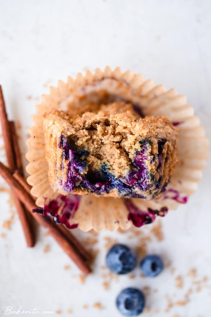 One blueberry muffin on it's side with a bite taken out to show the blueberries on the inside. On a white background and garnished with cinnamon sticks and fresh blueberries.