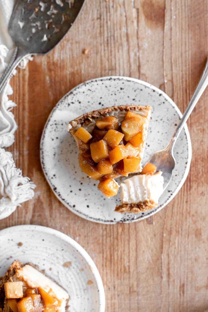 Slice of apple pie cheesecake with a fork bite taken out of it, photographed from above on a wooden board with a vintage cake server on the side.