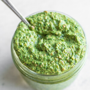 Power greens pesto in a small glass jar on a white background with a spoon coming out of the jar.