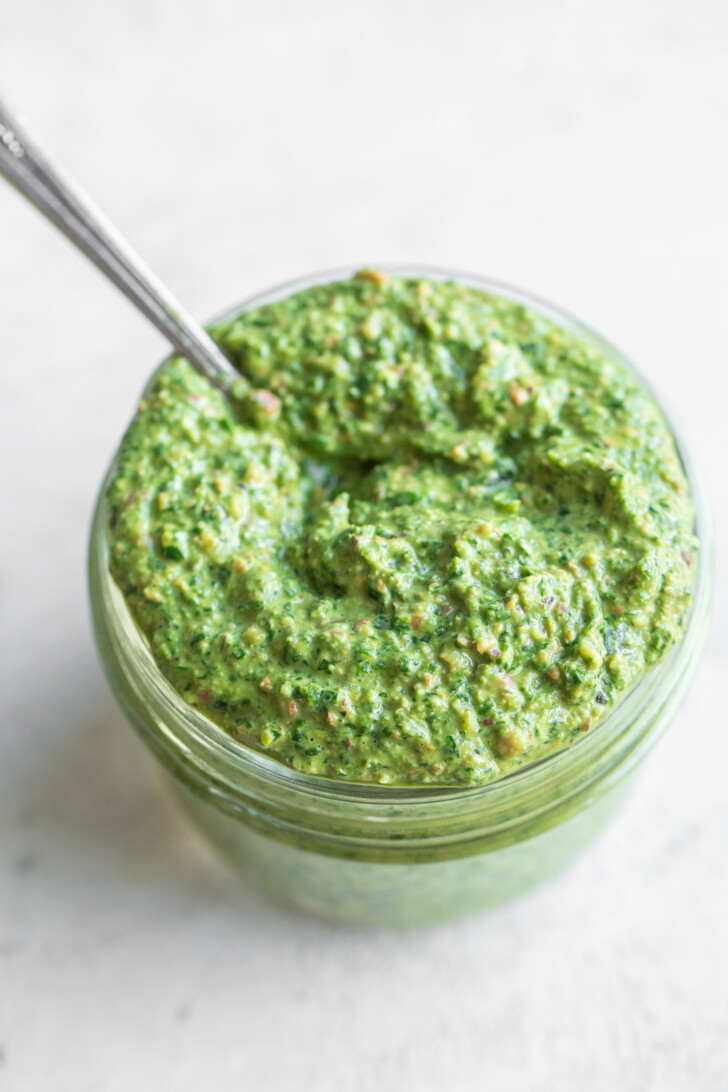 Power greens pesto in a small glass jar on a white background with a spoon coming out of the jar.